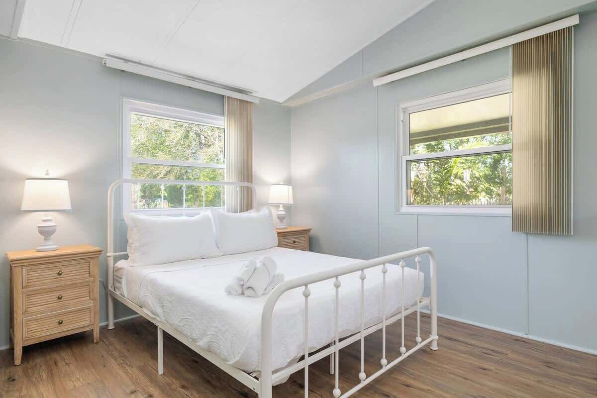 Un dormitorio con una cama de metal blanco, dos mesitas de noche con lámparas y ventanas amplias con cortinas.