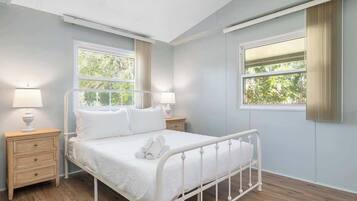 Un dormitorio con una cama de metal blanco, dos mesitas de noche con lámparas y ventanas amplias con cortinas.