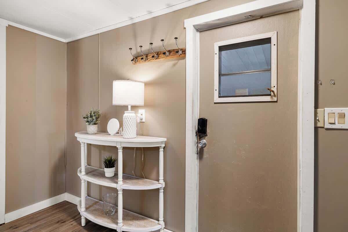 Ein Flur mit einer weißen Tür, einem Fenster, einem Kleiderständer, einem Tisch mit Lampe und einem Blumentopf.