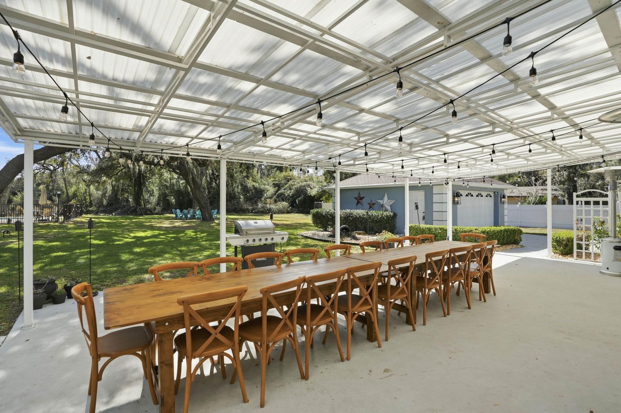 A covered patio with a long wooden table and chairs, a grill, and a view of a garden and house in the background.