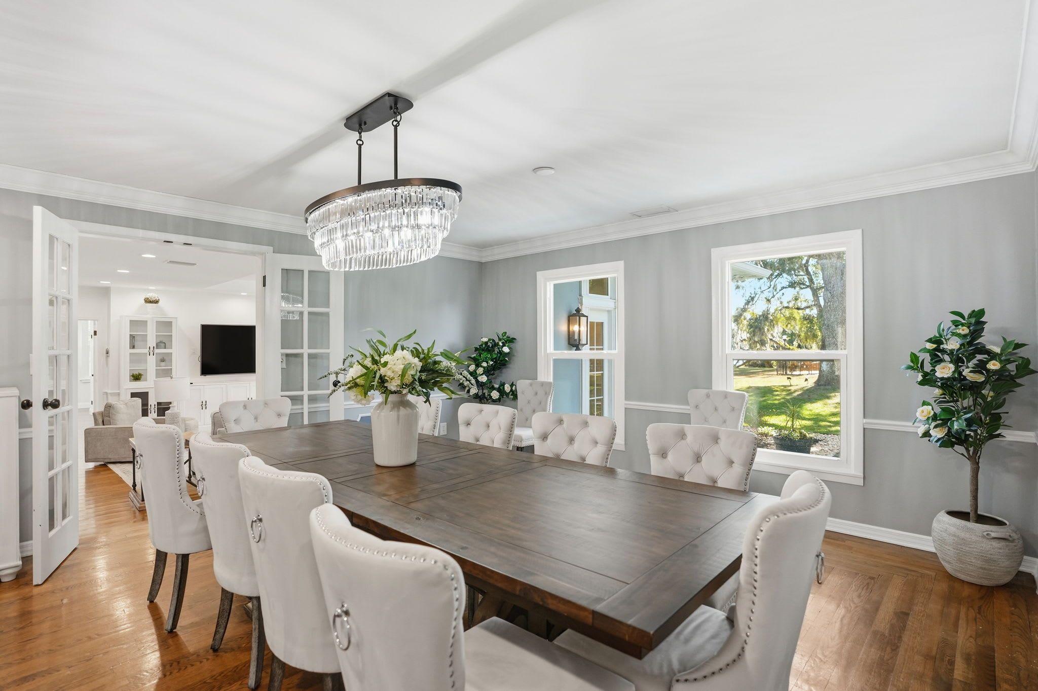 A spacious dining room with a large wooden table, white upholstered chairs, and a crystal chandelier.