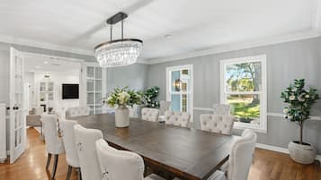 A spacious dining room with a large wooden table, white upholstered chairs, and a crystal chandelier.