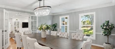 A spacious dining room with a large wooden table, white upholstered chairs, and a crystal chandelier.