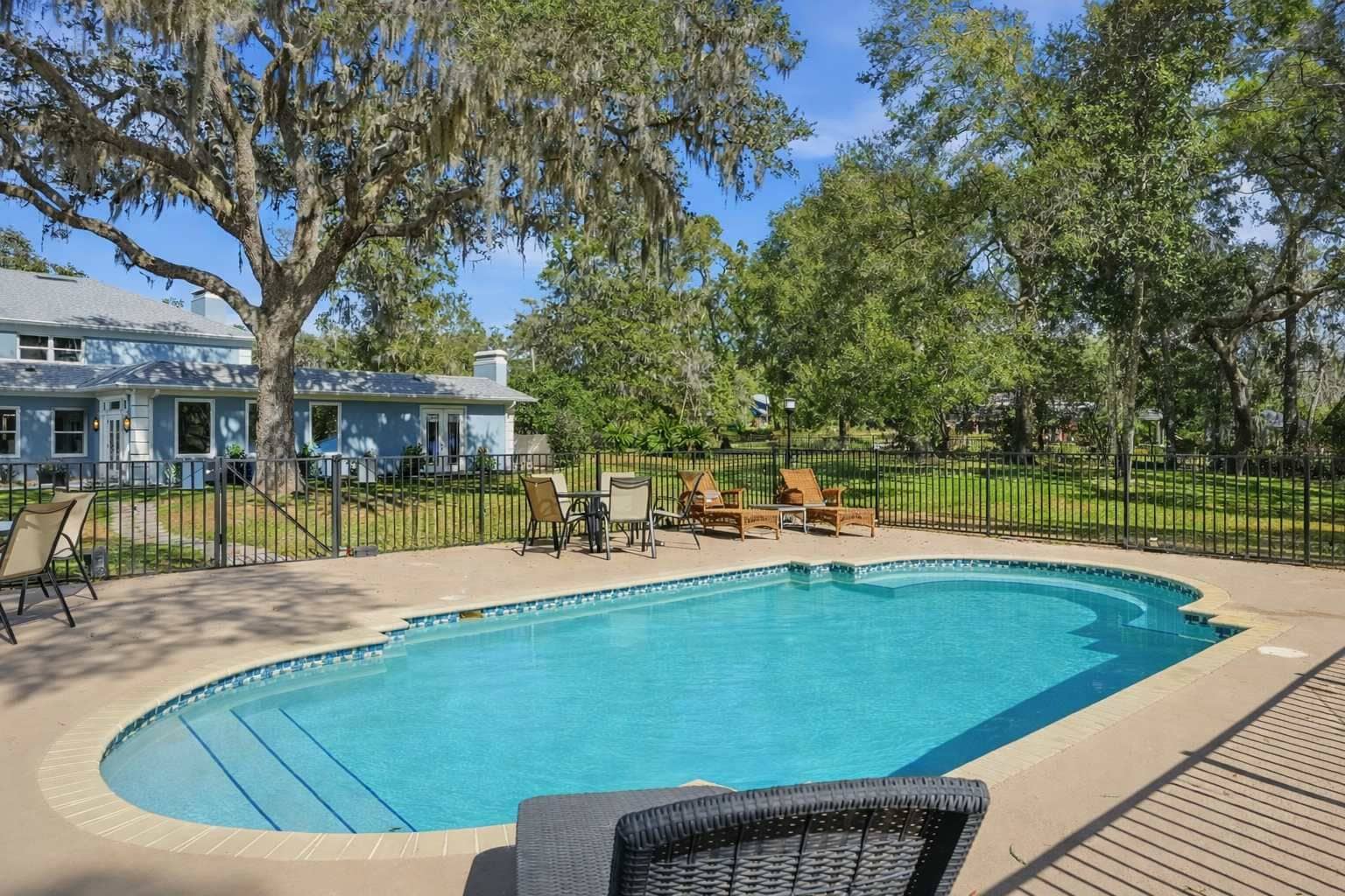 A swimming pool with lounge chairs and a fenced area, surrounded by trees and a house.