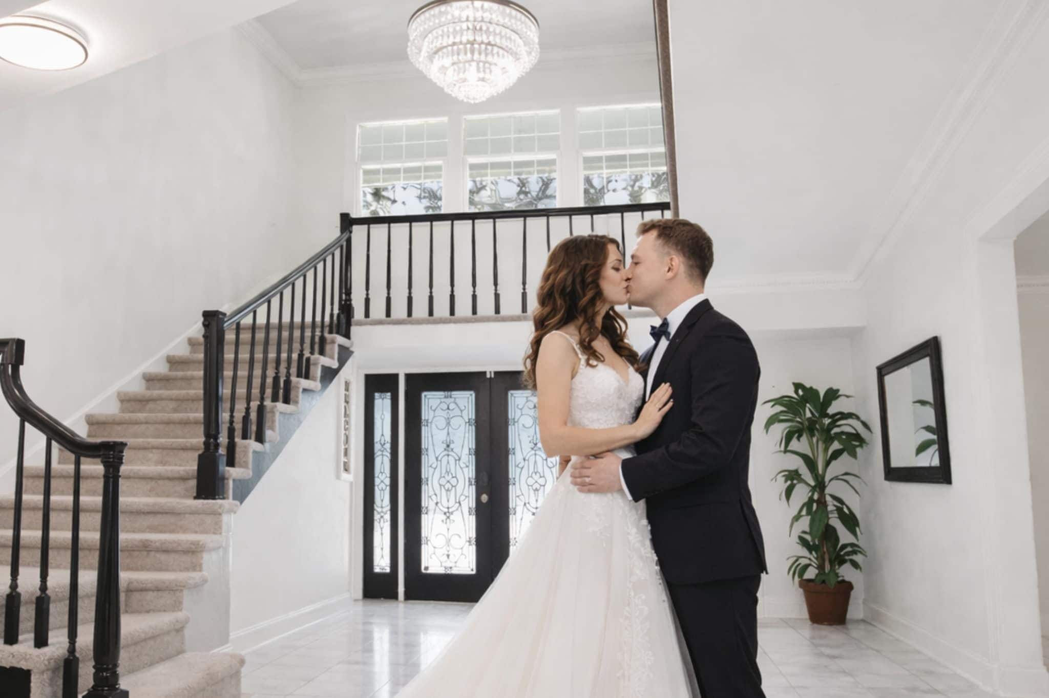 Una pareja se abraza en un vestíbulo amplio y luminoso, con una escalera imponente y un candelabro.