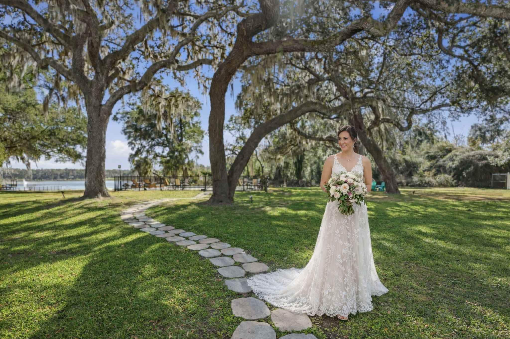 A wedding scene with a bride in a white dress standing on grass with a stone path and large trees in the background.