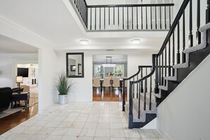A spacious living area with a high ceiling, a staircase with black railings, and a dining area in the background. - Executive 5 Bedroom Pool Home, Dade City! (Dade City)