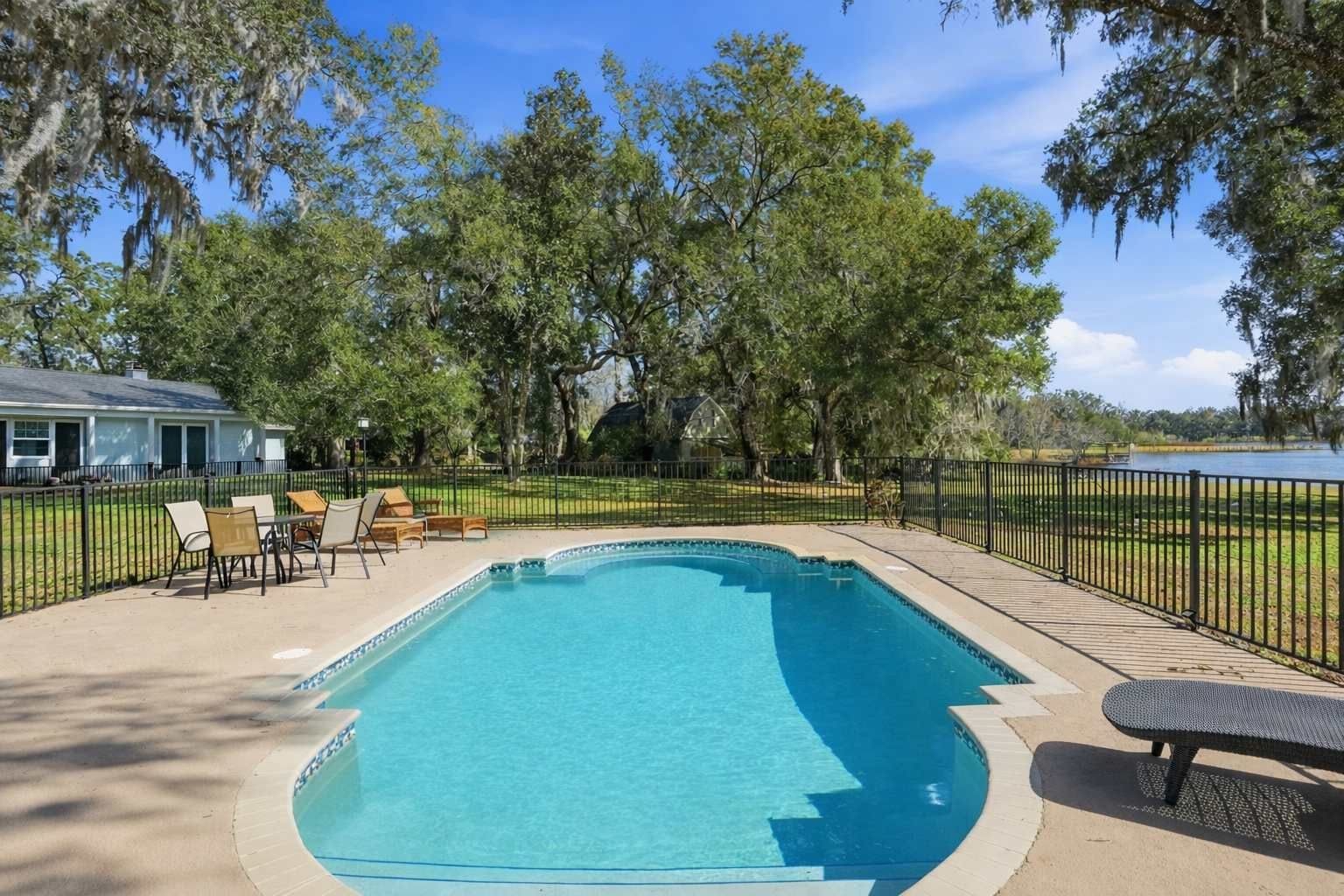 A rectangular swimming pool with a tiled deck, surrounded by a black metal fence, and shaded by trees.