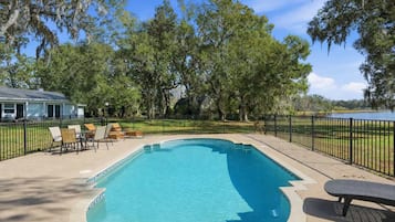 Una piscina rectangular con solárium de baldosas, rodeada por una valla de metal negro y protegida por árboles.