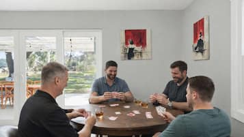 A group of four men playing cards at a round table in a room with two paintings on the walls and a view of the outdoors through a glass door.