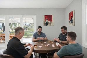 Un grupo de cuatro hombres jugando cartas alrededor de una mesa redonda en una habitación con dos cuadros en las paredes y vistas al exterior a través de un ventanal.