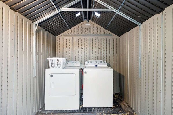 A laundry room with two washing machines and a dryer, metal walls, and a sloped ceiling with skylights.