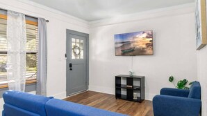 A living room with a grey door, a blue sofa, a blue armchair, a black TV stand, and a painting of a sunset over water. - Charming 1-bedroom Home in Lakeland! (Lakeland)