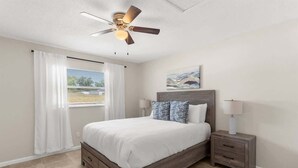 Quarto com cama, mesas de cabeceira, ventoinha no teto e janela com cortinas.