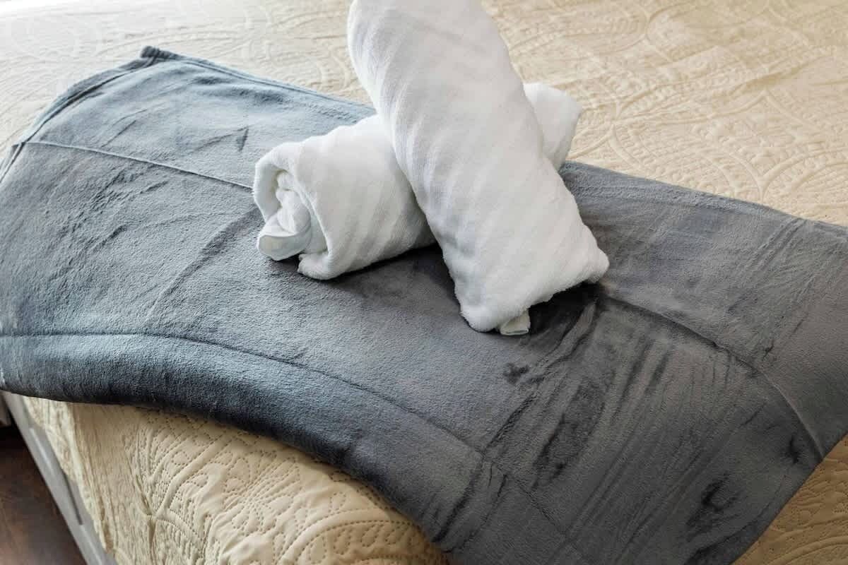 Una cama con un almohadón gris y una toalla blanca doblada.