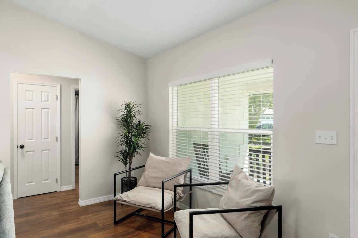 Una habitación con piso de madera, una puerta blanca, una ventana con persianas y dos sillas con cojines.