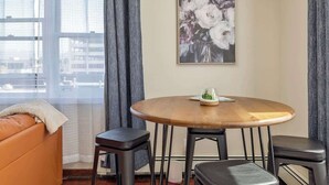 A modern room with a round wooden table, two black bar stools, a beige sofa, and a large floral painting on the wall. - Historical Art Deco Central Downtown - Sleeps 4 (Anchorage)