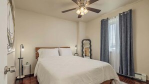 A bedroom with a bed, a ceiling fan, a nightstand, a mirror, and a window with curtains. - Historical Art Deco Central Downtown - Sleeps 4 (Anchorage)