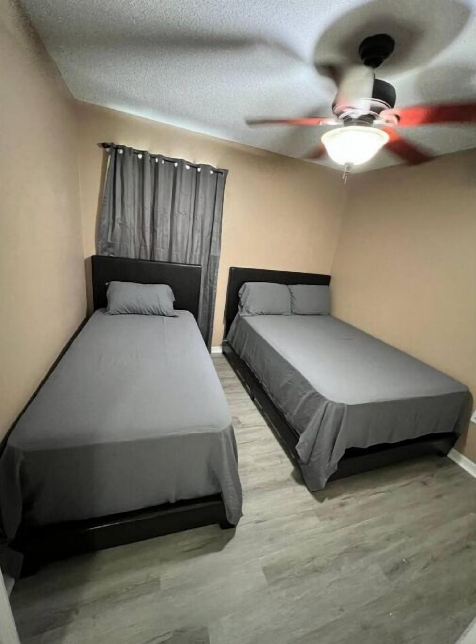 Una habitación con dos camas individuales, un ventilador de techo y una ventana con cortinas.