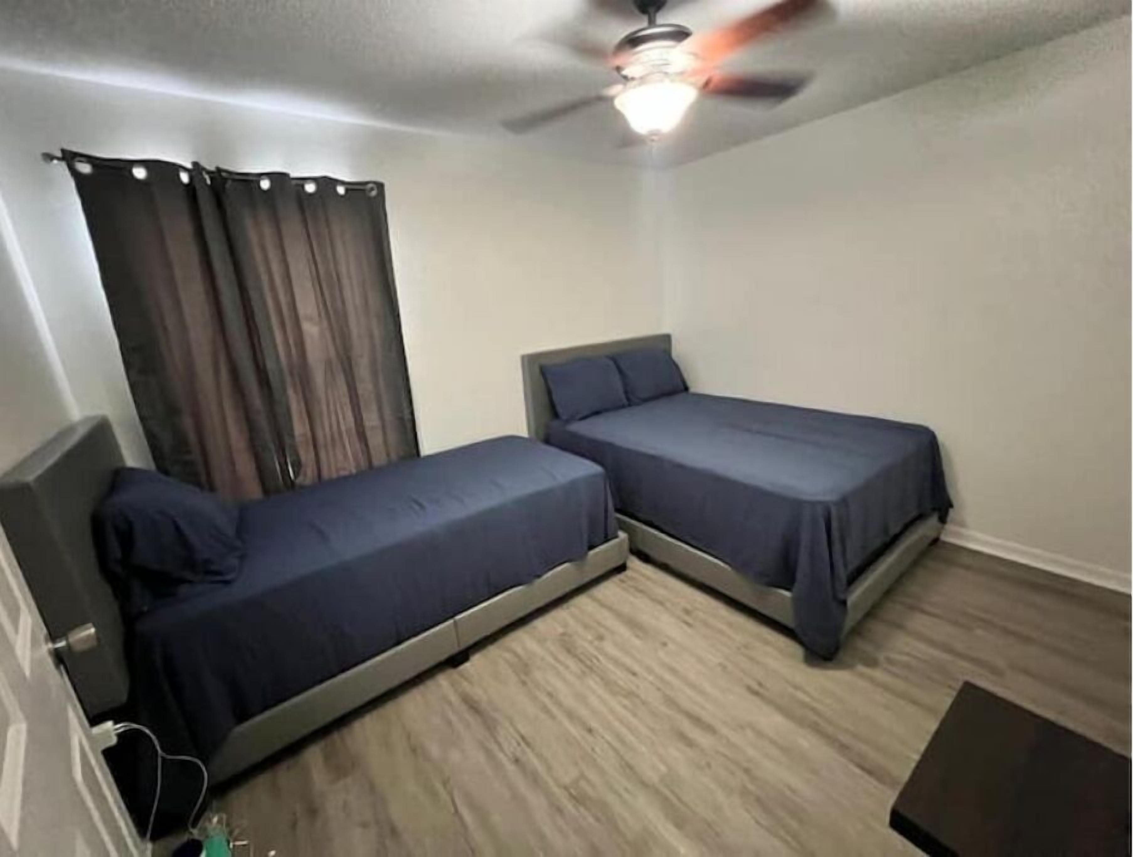 A small, single-room hotel room with two single beds, a ceiling fan, and a window with curtains.