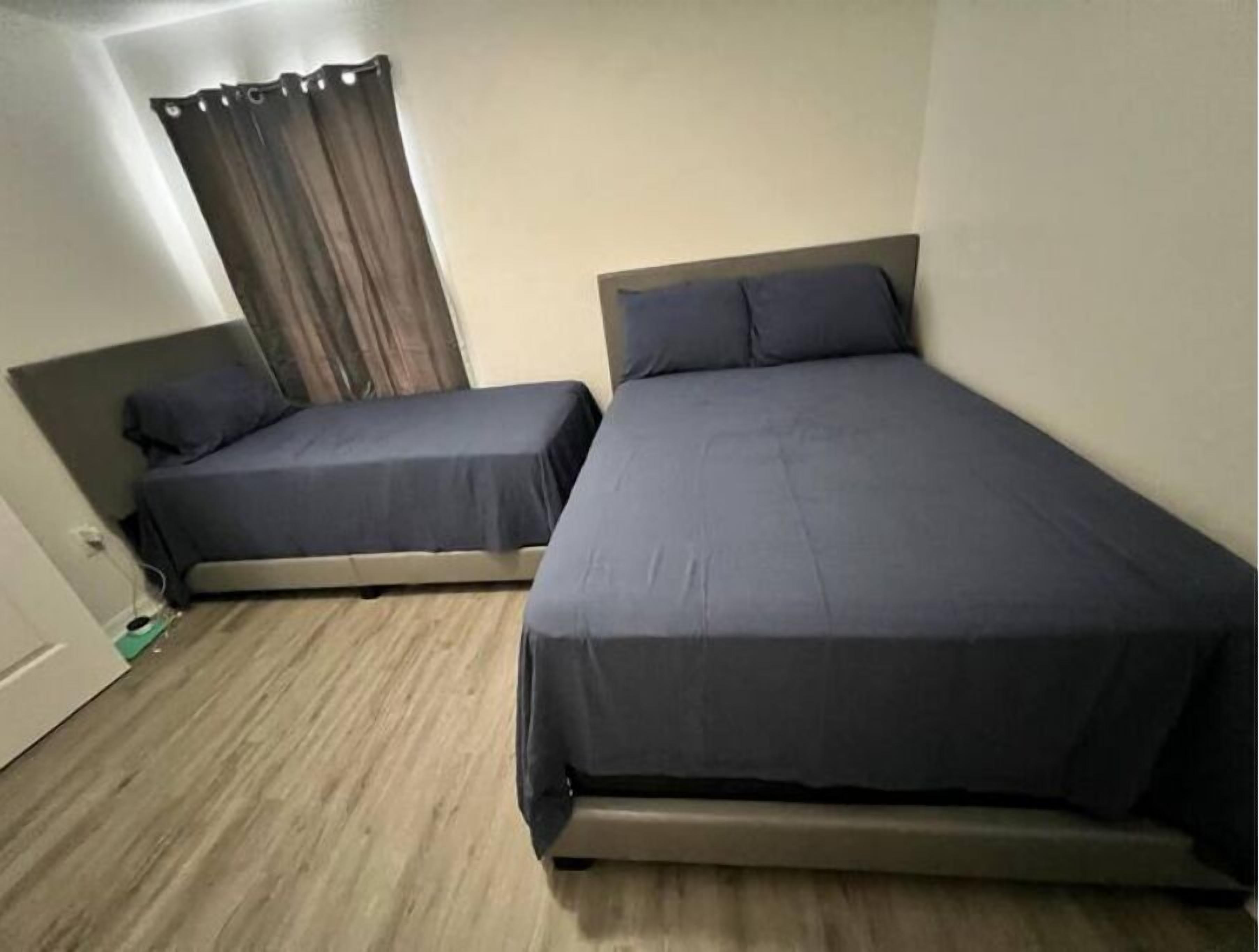 Ein kleines Hotelzimmer mit einem Einzelbett, zwei Betten, Holzboden und einem Vorhang am Fenster.
