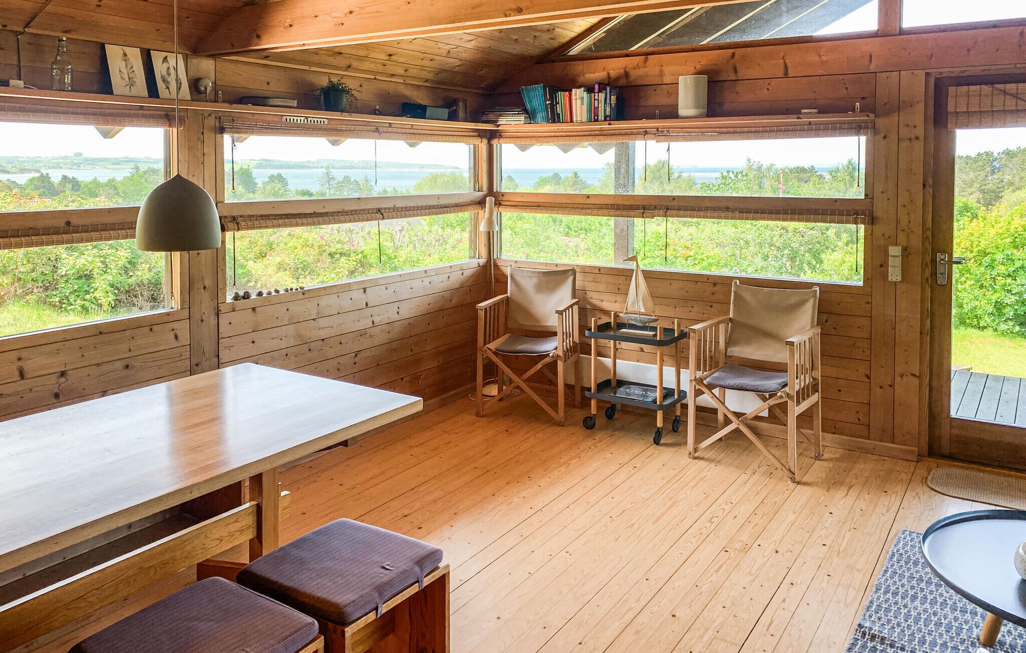 A wooden cabin interior with a dining table, two chairs, a small cart, and large windows offering a view of a lake and greenery.