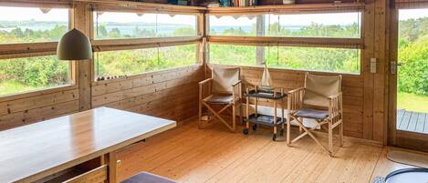 A wooden cabin interior with a dining table, two chairs, a small cart, and large windows offering a view of a lake and greenery.