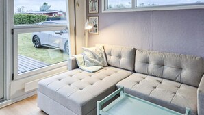 A modern living room with a light grey sofa, a small table, and a window with a view of a car and greenery outside.