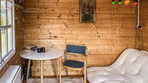 A cozy cabin room with a wooden interior, a round table with chairs, a bed, and a window with curtains.
