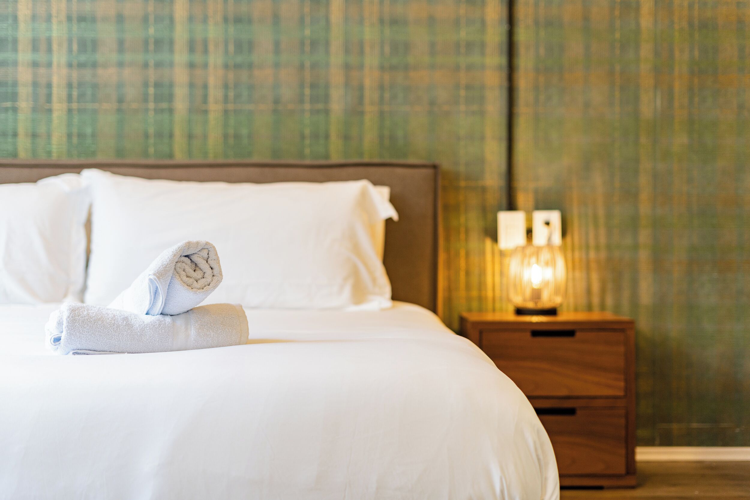 A neatly made bed with a rolled towel and pillows, a wooden nightstand with a lamp, and a patterned wall in the background.