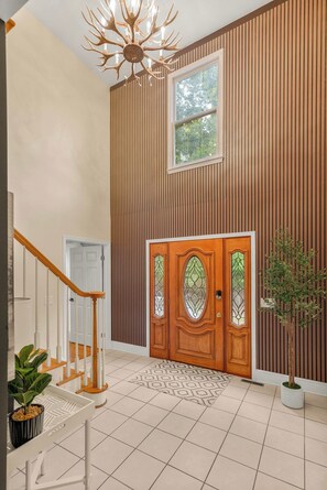 A modern interior with a wooden-paneled wall, a large wooden door with glass panels, a staircase with white railings, and a chandelier.