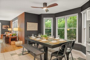 Zona pranzo con tavolo scuro apparecchiato per quattro persone, ventilatore a soffitto e ampie finestre con vista sul verde.