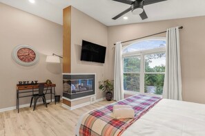 A bedroom with a bed, a desk, a television, a fireplace, and a large window with curtains.