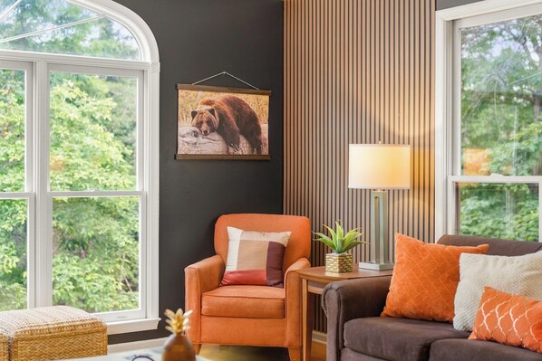 A room with a large window, a chair, a sofa, a lamp, and a picture of a bear on the wall.