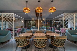 Een moderne zithoek met ronde tafels en rieten stoelen, met een centraal decoratief element en verlichting aan het plafond.