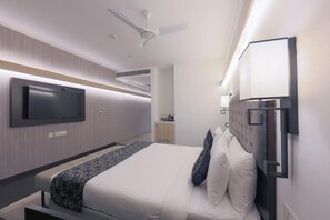 Una habitación de hotel moderna con una cama grande, un televisor de pantalla plana montado en la pared, un ventilador de techo y una mesita de noche con lámpara.