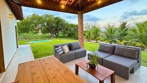 An outdoor patio with a wooden table and two grey sofas, a brick pillar, and a view of a swimming pool and greenery. - Casa Da Costa (Aracati)