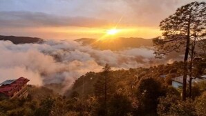 Un paisaje montañoso con amanecer, nubes bajo los árboles y edificios en primer plano.