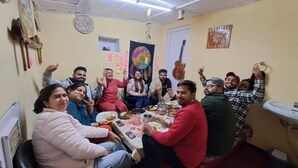 A group of people seated around a table in a room with a white ceiling, a guitar on the wall, and a sink to the right. - Delicate camp C (Kiar)