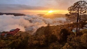 Eine Berglandschaft bei Sonnenaufgang, Wolken unter den Baumwipfeln und Gebäude im Vordergrund.