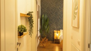 A narrow hallway with a wooden floor, a bed with a nightstand, a lamp, and a large plant. - Prime Living - [Apartment GOLD] on the Navigli (Milano)
