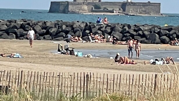En strand med folk, der solbader og svømmer, et fyrtårn i baggrunden og en stenmole.