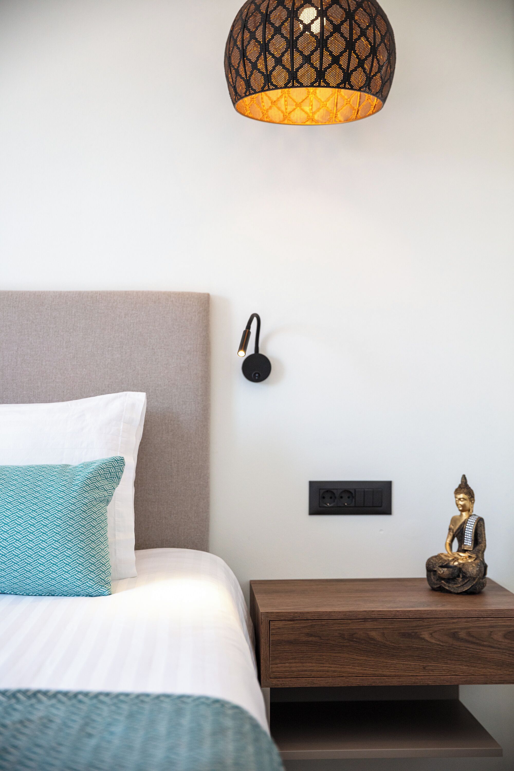 A neatly made bed with white and teal pillows, a wooden nightstand with a Buddha statue, a wall-mounted light fixture, and a power outlet.