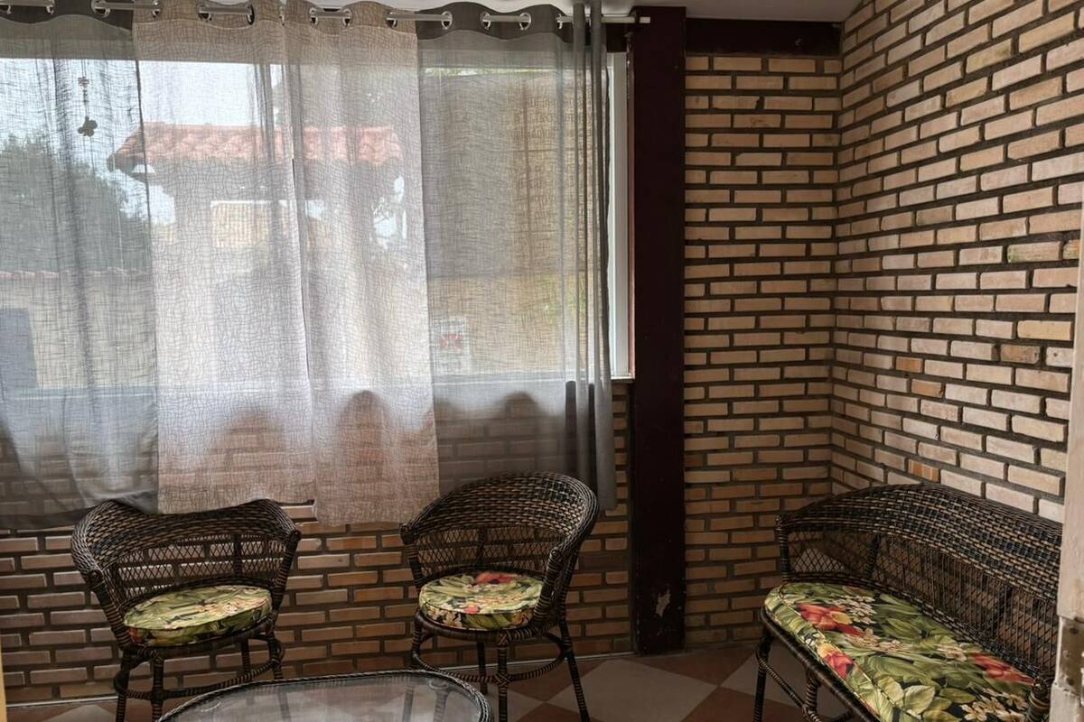 Ett hörnrum med en tegelvägg, två flätade fåtöljer med blommiga kuddar, ett runt glasbord och ett fönster med en tunn, genomskinlig gardin.