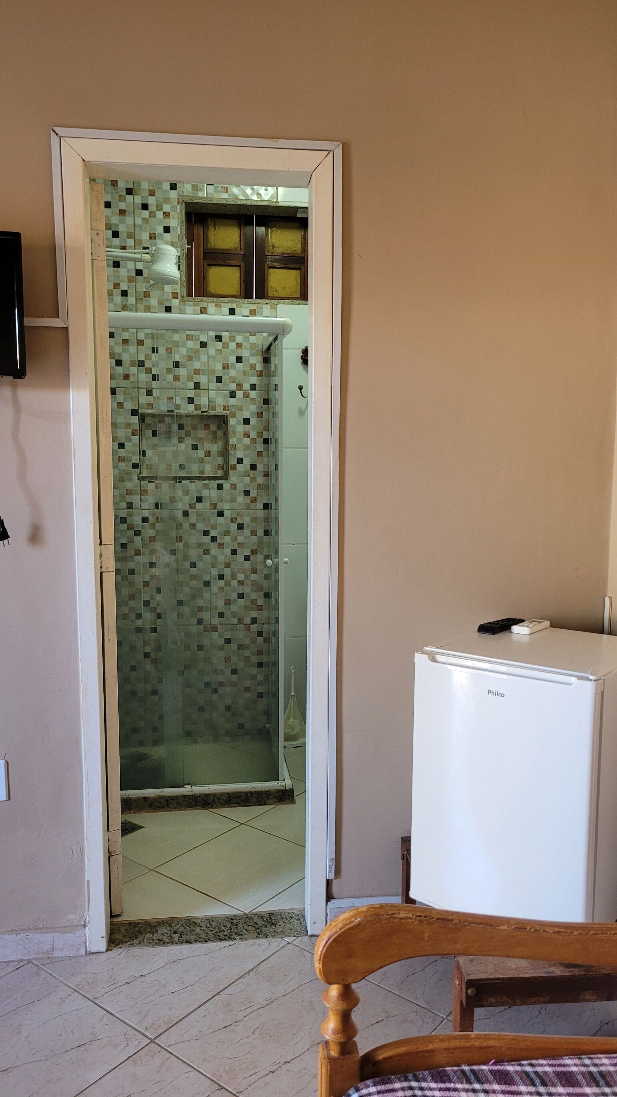 Quarto com chuveiro azulejado, geladeira branca e cadeira de madeira.