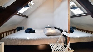 Une petite chambre mansardée, chaleureuse et accueillante, avec un plafond en pente, un lit recouvert d’une couvre-lit à motifs, deux tables de chevet avec des lampes et une chaise en bois.