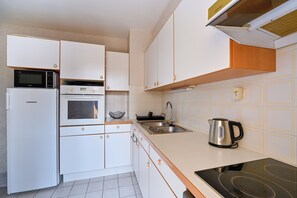 Cocina compacta con armarios blancos, microondas, horno, fregadero y hervidor.