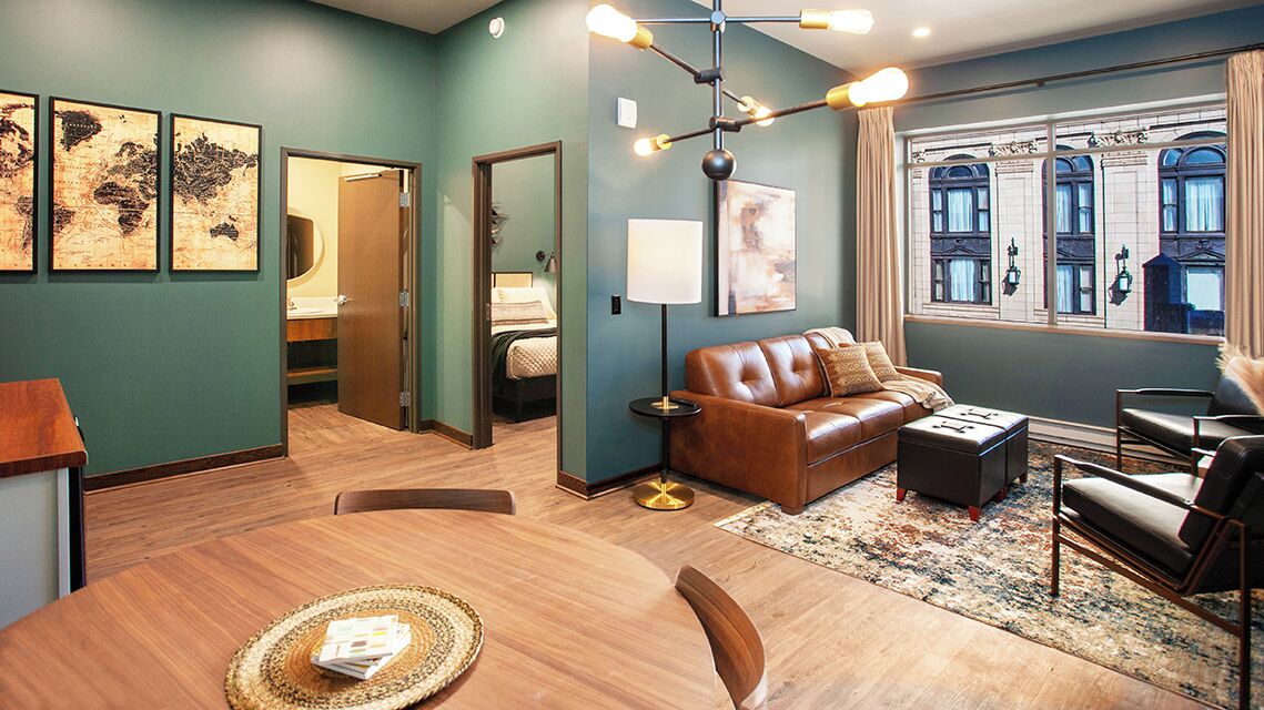 A modern hotel room with a wooden dining table, leather sofa, and a large window with a view of historic buildings.