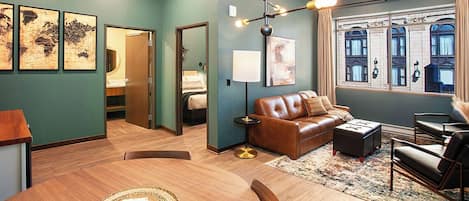 A modern hotel room with a wooden dining table, leather sofa, and a large window with a view of historic buildings.