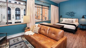 A modern hotel room with a large window, a brown leather sofa, a bed with a white headboard, and a small table. - The Intrepid (Duluth)